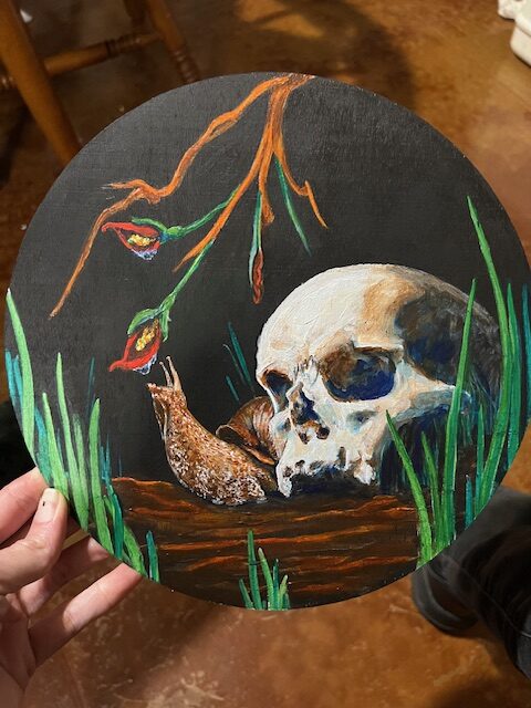 A painting of skull with a snail reaching up for a drink of water that has collected on a flower.